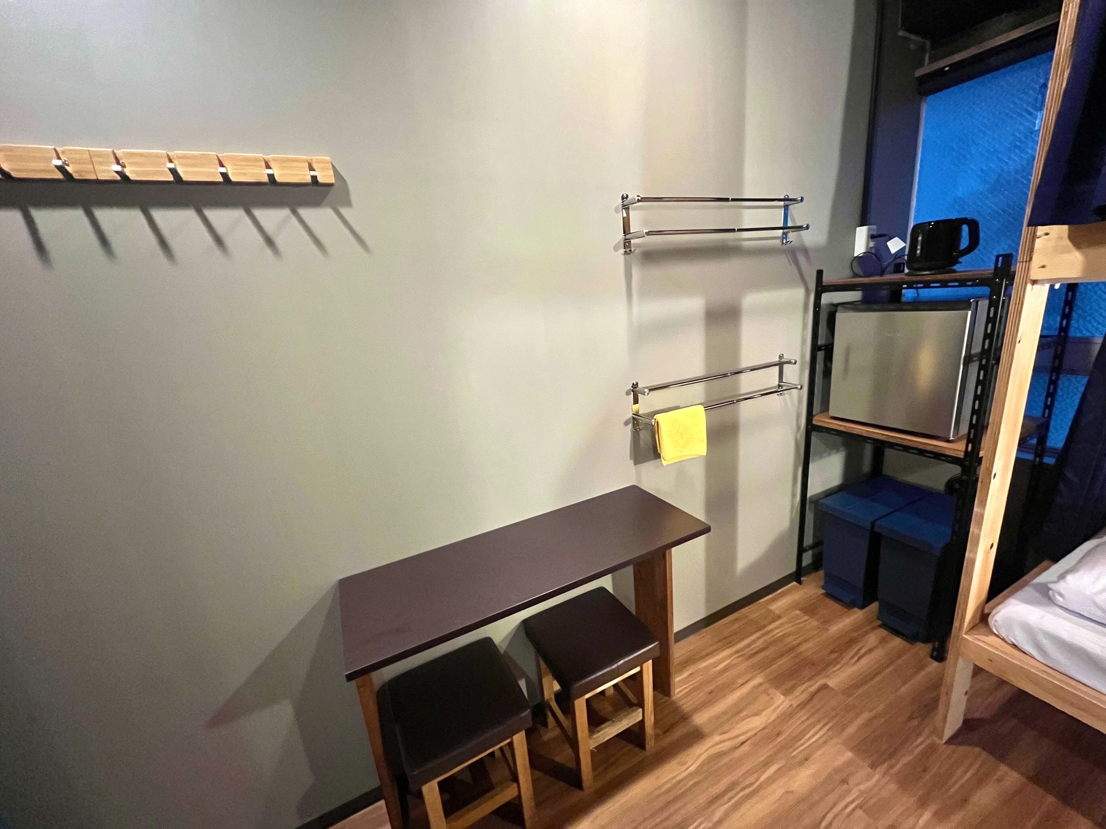 A compact room with a wooden desk, two chairs, a small stool, a metal towel rack, and a bunk bed with a blue window.