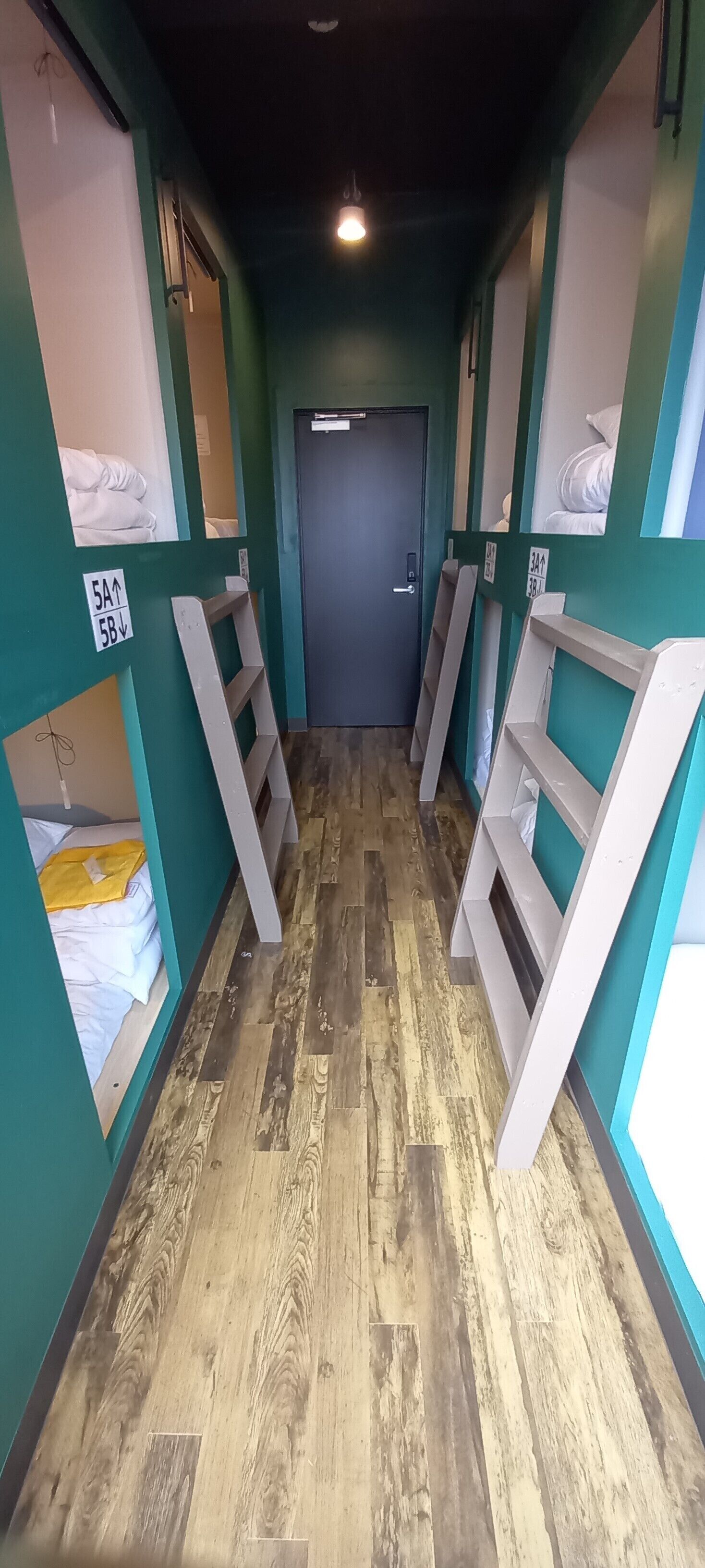 A hallway with wooden flooring, green walls, and beds with white bedding. There are signs indicating room numbers and directions.
