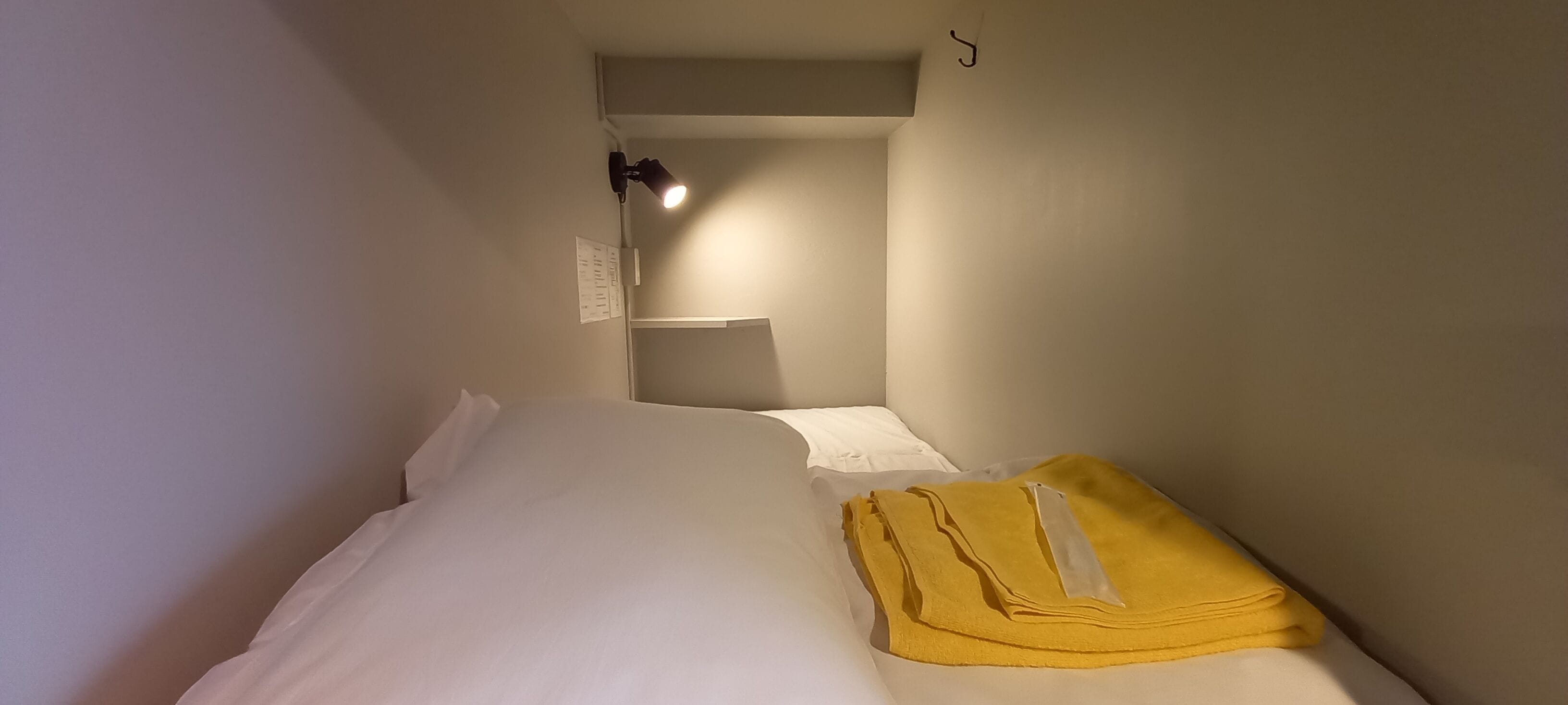 A small, narrow room with a bed, a yellow towel, a white pillow, and a wall-mounted light fixture.