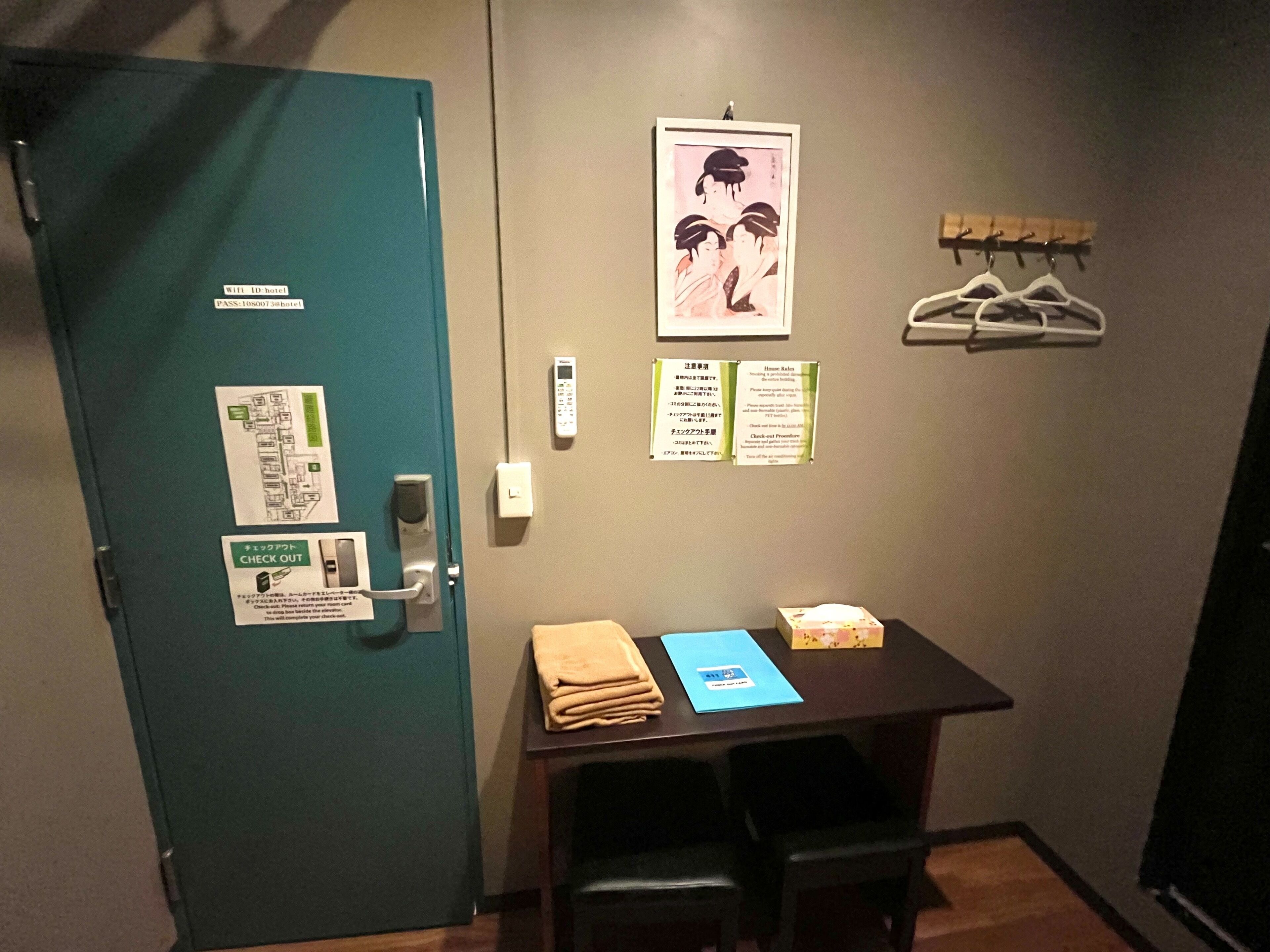 A hotel room with a teal door, a check-out sign, a map, a digital lock, a table with a towel, a blue folder, and a small box.