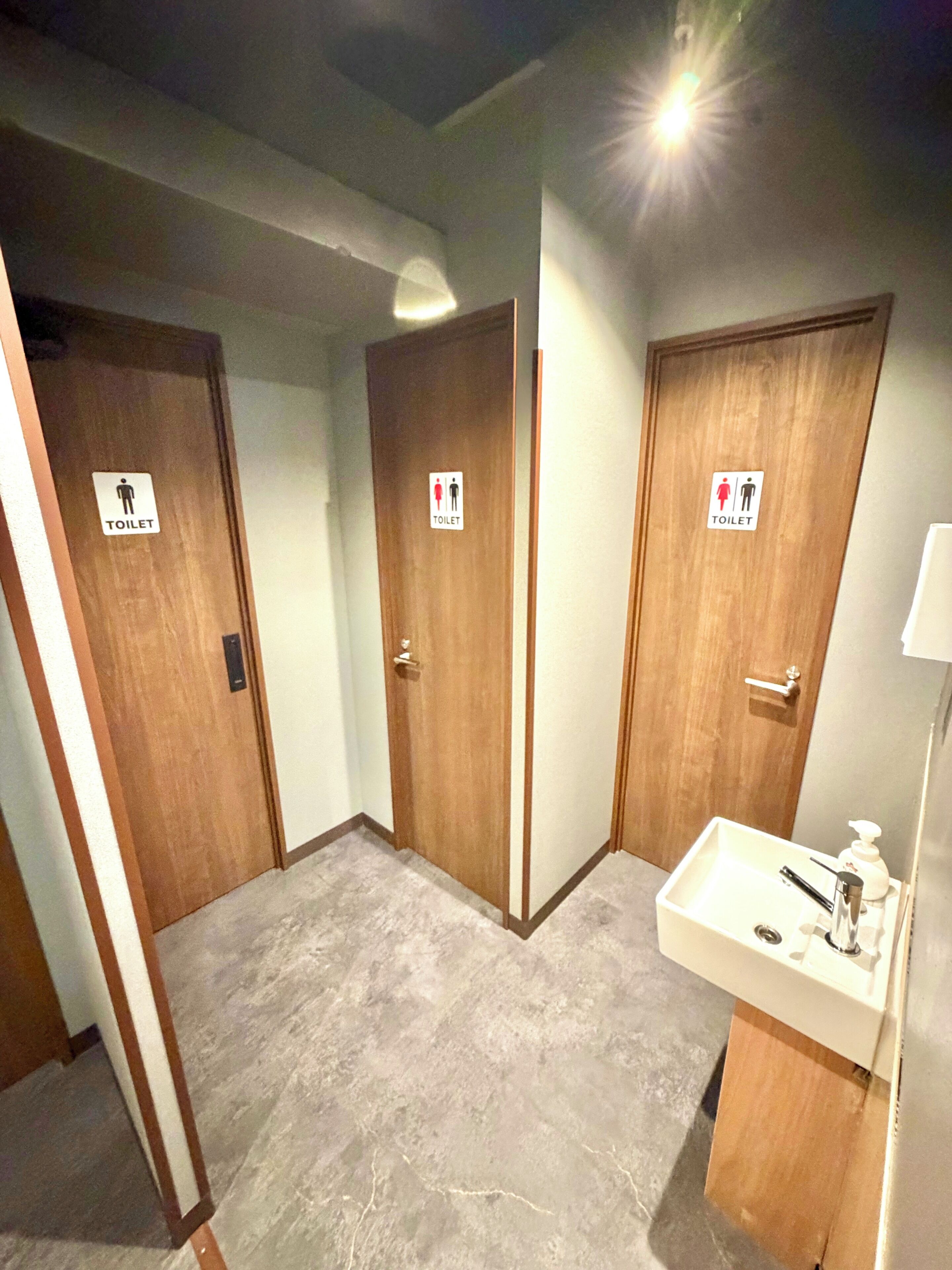 A hotel bathroom with three wooden doors, each marked with a toilet sign, and a sink with a mirror and soap dispenser.