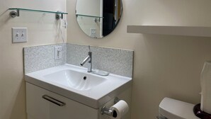 Een badkamer met een witte wastafel, een ronde spiegel, een glazen plank boven de wastafel en een toilet met een papieren handdoekhouder.
