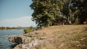 Un chalet rustique au bord d’un lac, avec une colline herbeuse et des arbres.