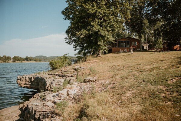 Ein rustikales Blockhaus an einem See, mit einer grasbewachsenen Hügelkuppe und Bäumen.