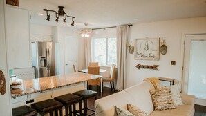 Une cuisine moderne dotée d’un îlot central, de tabourets de bar, d’un coin repas avec une chaise, et d’un salon avec un canapé et des coussins décoratifs.