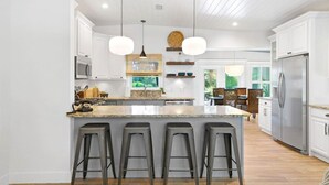 A modern kitchen with white cabinetry, a central island with bar stools, pendant lighting, and a view to a dining area. - Villa Solano| Holiday deals, book now! (Downtown St. Augustine)