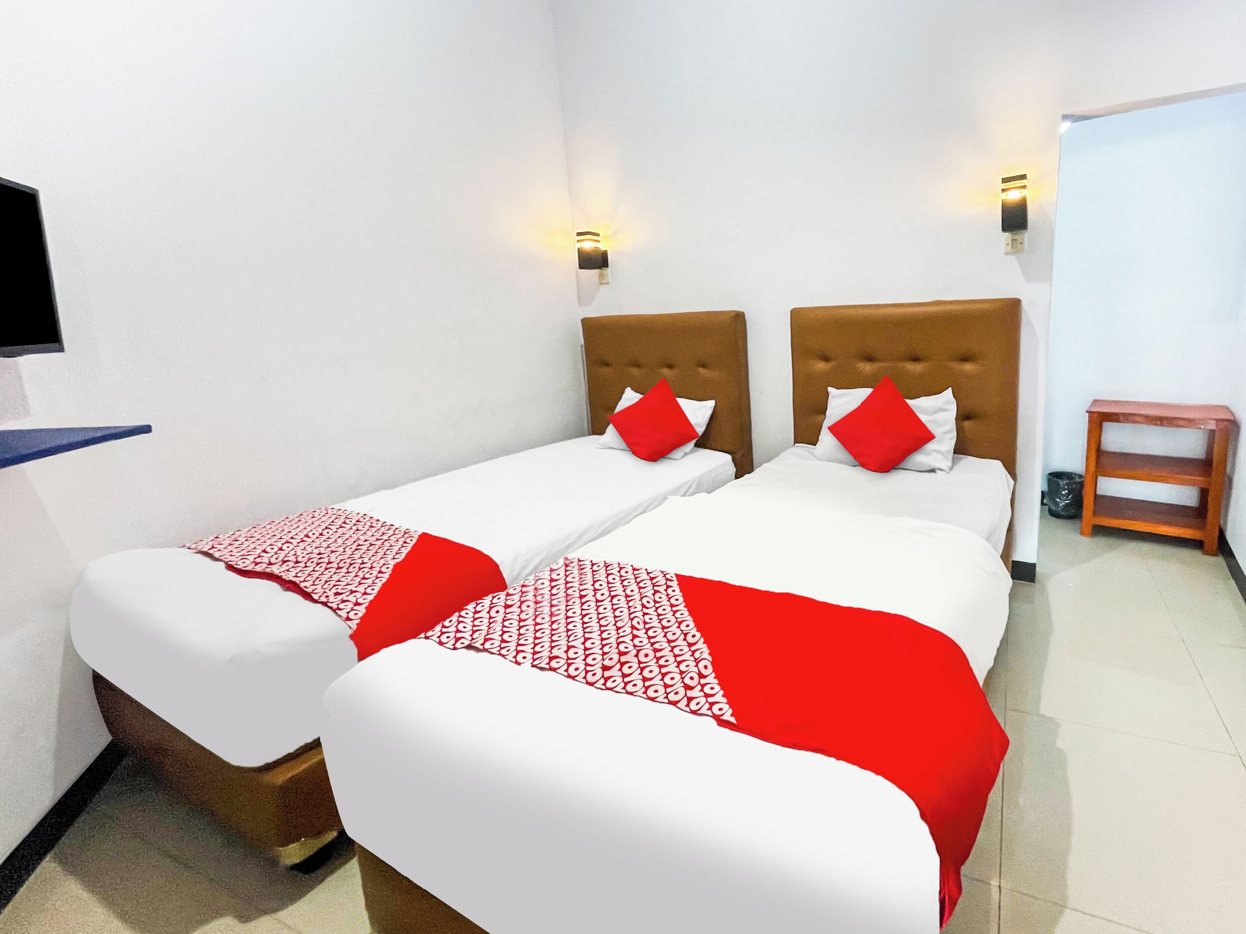 Ein Hotelzimmer mit zwei Betten, jeweils mit Kopfteil, einem kleinen Nachttisch aus Holz und einem an der Wand montierten Fernseher.