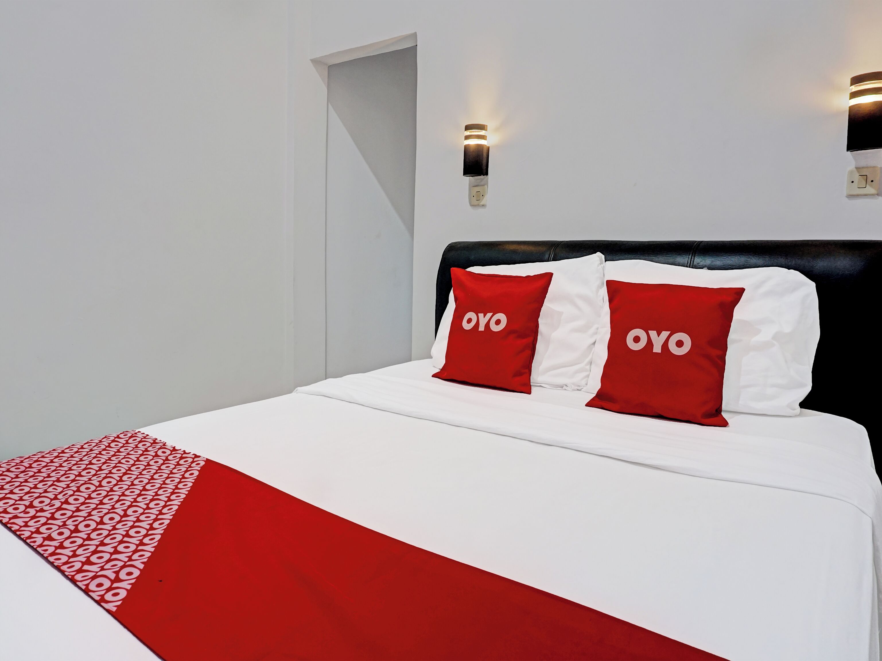 Ein Bett mit weißer Bettwäsche und zwei roten Kopfkissen mit 'OYO'-Aufschrift, ein schwarzer Kopfteil und zwei an der Wand befestigte Lampen.