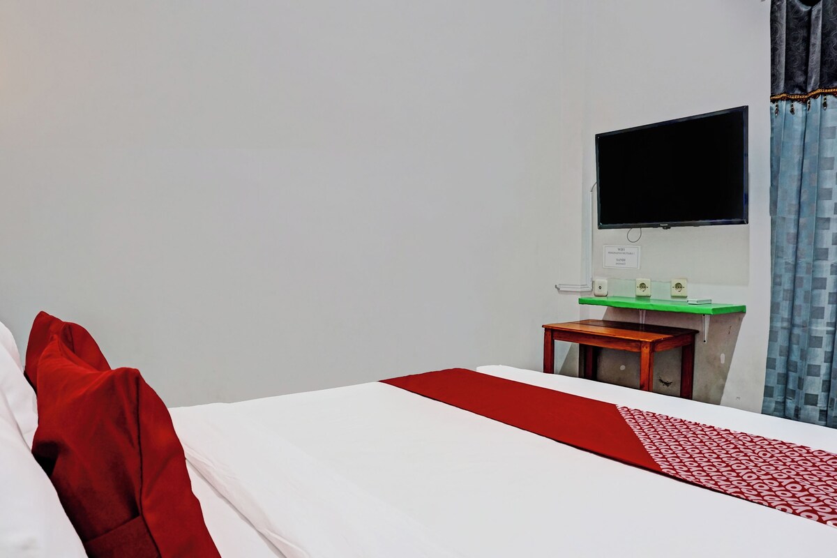 A hotel room with a bed, a small wooden table, a green countertop, a TV mounted on the wall, and a window with curtains.