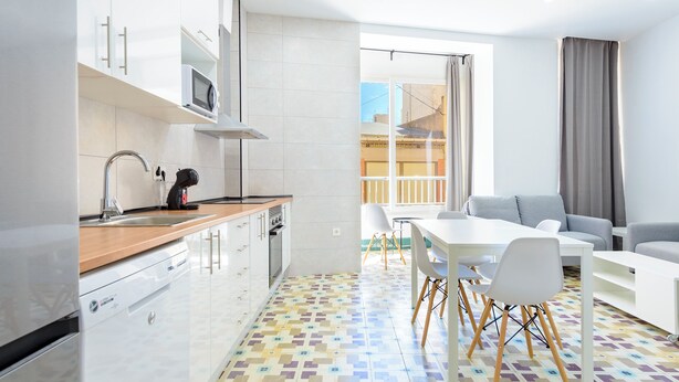 A modern kitchen with white cabinets, a microwave, and a coffee machine. There is a dining area with a white table and chairs, and a gray sofa. The floor has a colorful geometric pattern. -  Bieti Castellon (Castelló de la Plana)