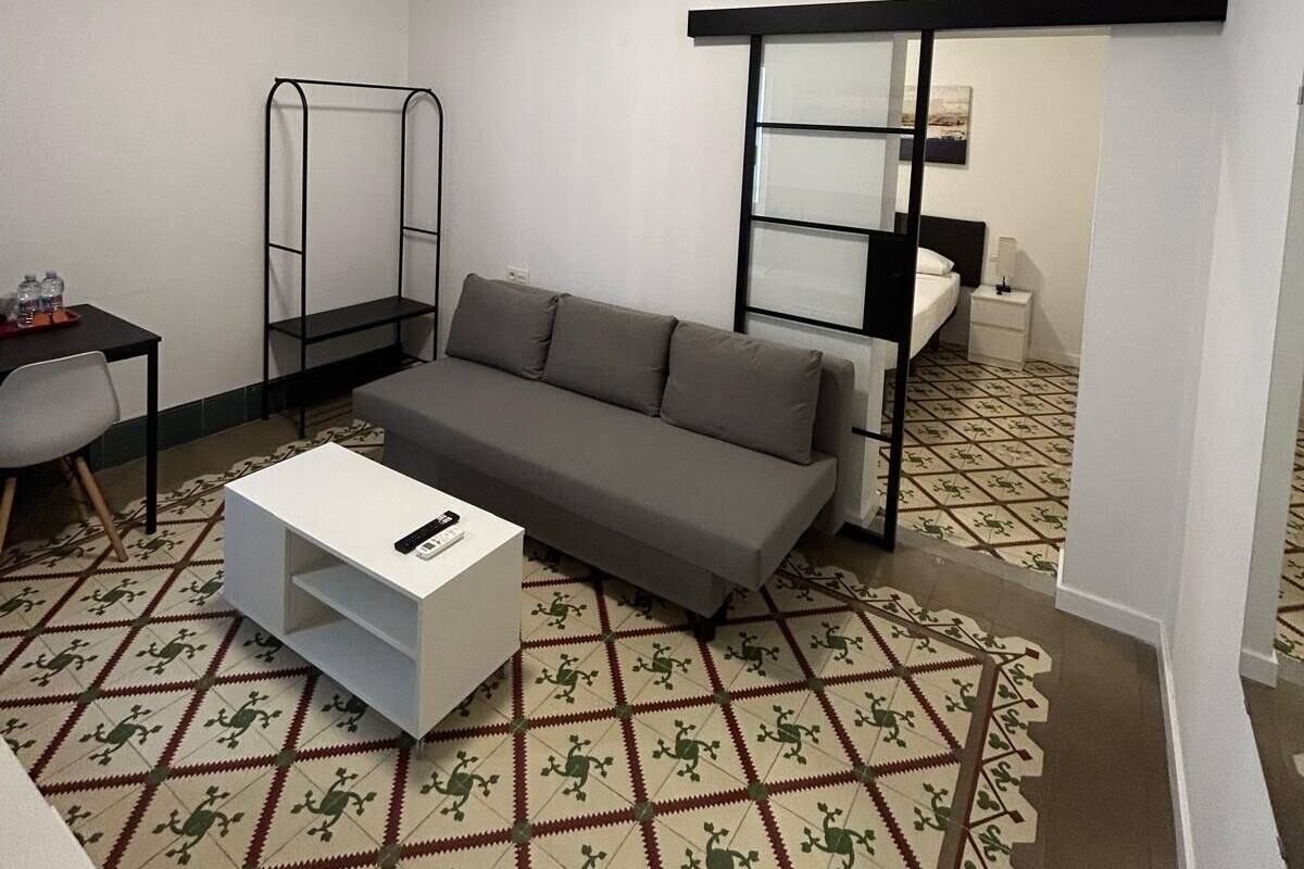 Un espace de vie compact comprenant un canapé gris, une table basse blanche, un petit bureau avec une chaise et un sol à motifs.