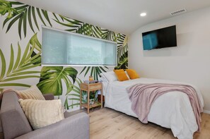 A bedroom with a bed, a TV, a window with blinds, a sofa, and a wall with a tropical leaf pattern.