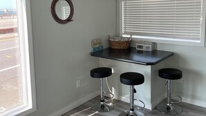 A kitchen with a dark countertop, three bar stools, a round mirror, and a window with blinds. - Ocean views from every window.  5min walk to the quiet end of the beach! (Hampton)