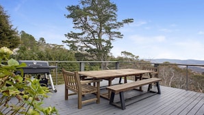 Eine Terrasss mit Holztisch und -bänken, einem Grill und Blick auf Bäume und Hügel.