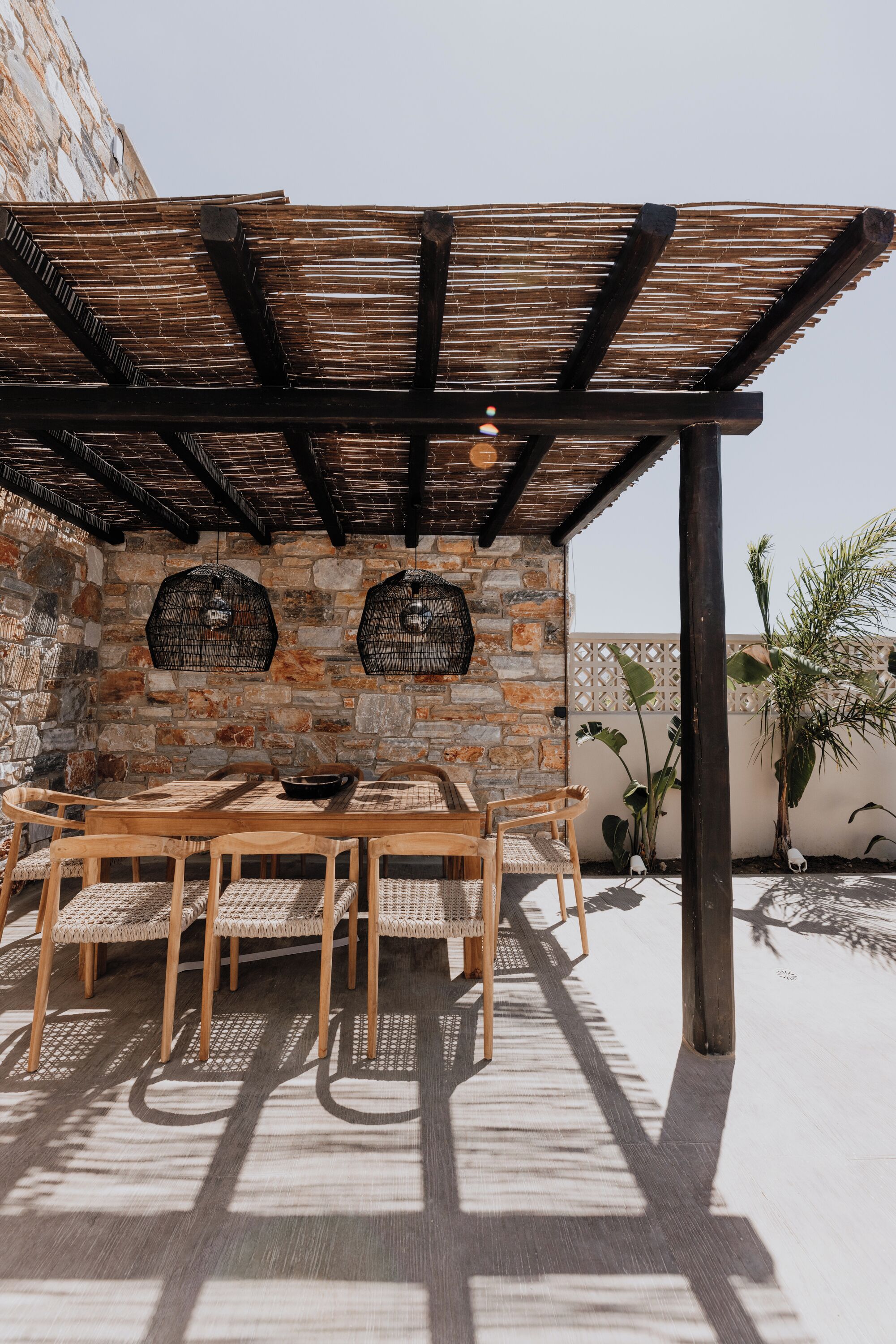 A covered outdoor dining area with a stone wall, wooden table and chairs, and hanging birdcages.