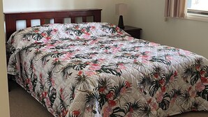Een slaapkamer met een groot bed, een nachtkastje, een lamp, een lijst met een afbeelding aan de muur en een raam met gordijnen.