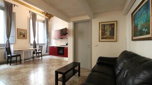 Un salon avec un canapé en cuir noir, une table basse en bois et un coin repas comprenant une table et des chaises. On peut apercevoir la cuisine à travers une porte ouverte.