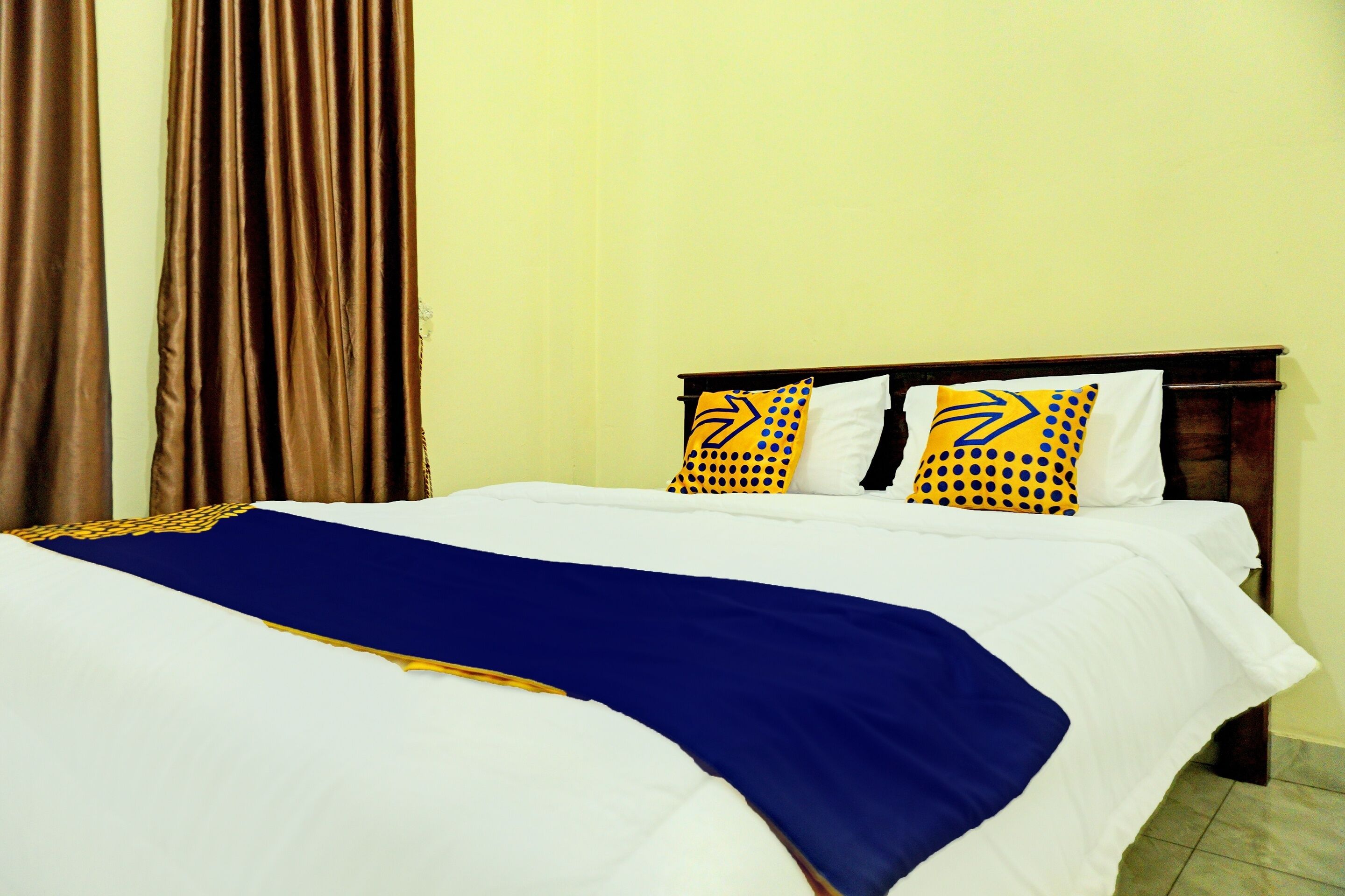 Una cama bien hecha con ropa de cama blanca y azul, dos almohadas con diseños amarillos y negros, y una cortina marrón.