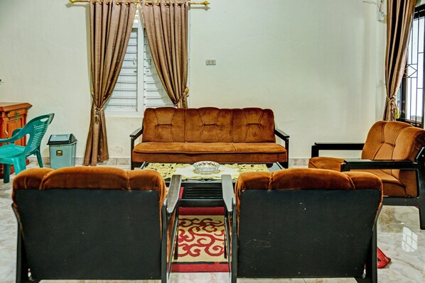 A living room with a brown sofa, two black chairs, a small table, and a window with curtains.