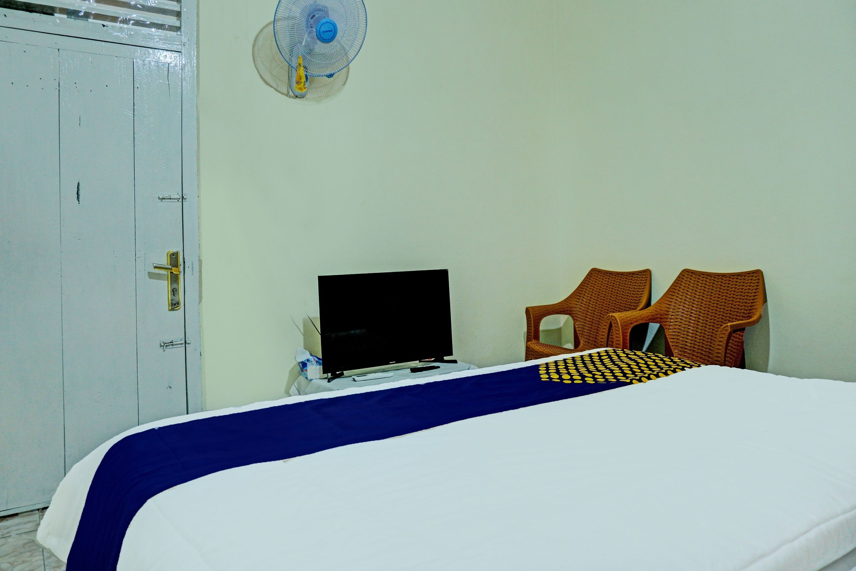 A room with a bed, a desk with a laptop, two wicker chairs, a white wardrobe, and a wall-mounted fan.