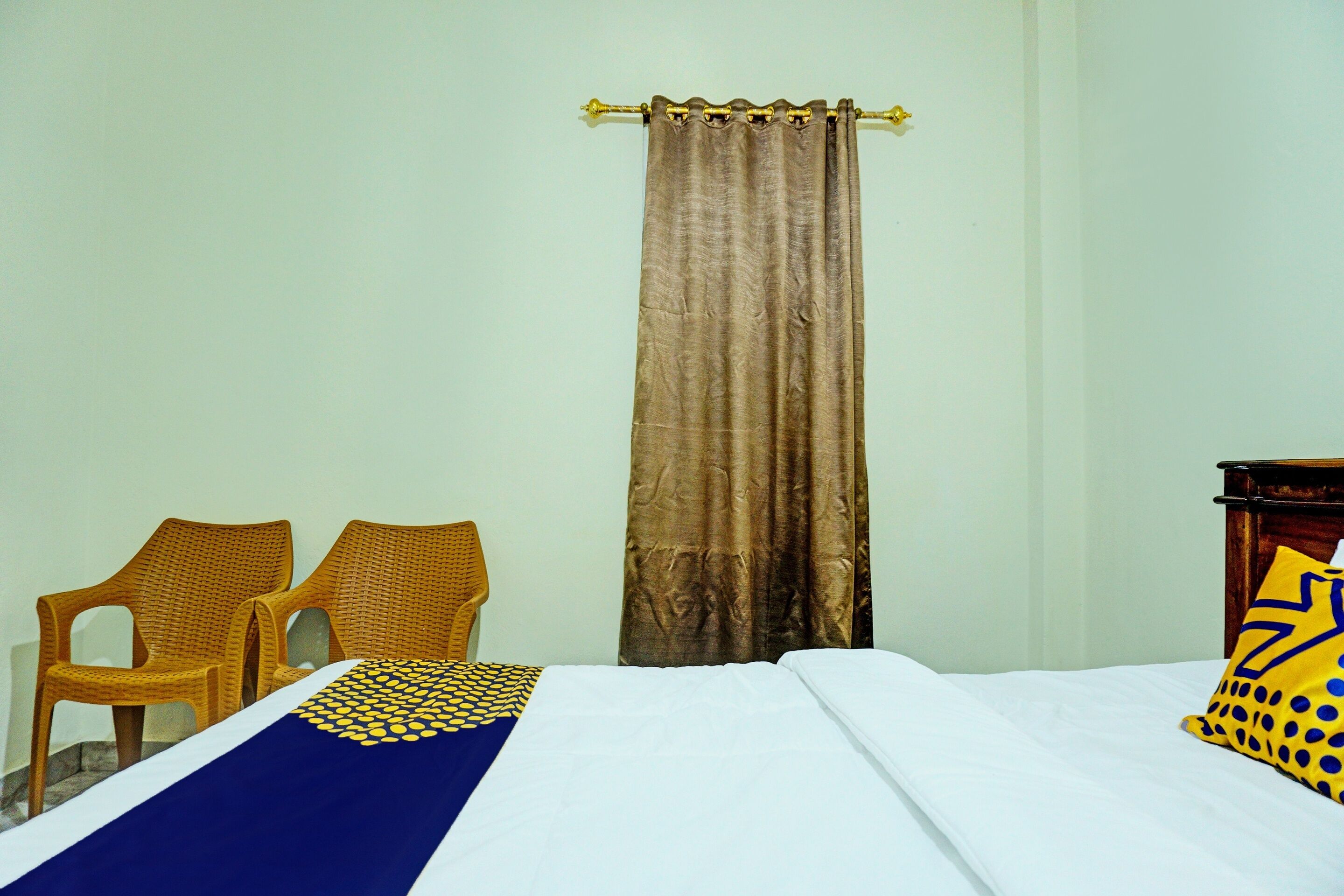 A room with a bed, two wicker chairs, a curtain, and a wooden nightstand with a pillow.