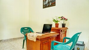 A room with a wooden desk, two green plastic chairs, a laptop, a potted plant, and a trash bin.