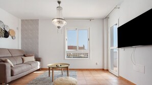 A living room with a sofa, a round ottoman, a small table, a chandelier, and a TV mounted on the wall. - Luxury Ocean View holiday home with sea view and shared pool (Corralejo)