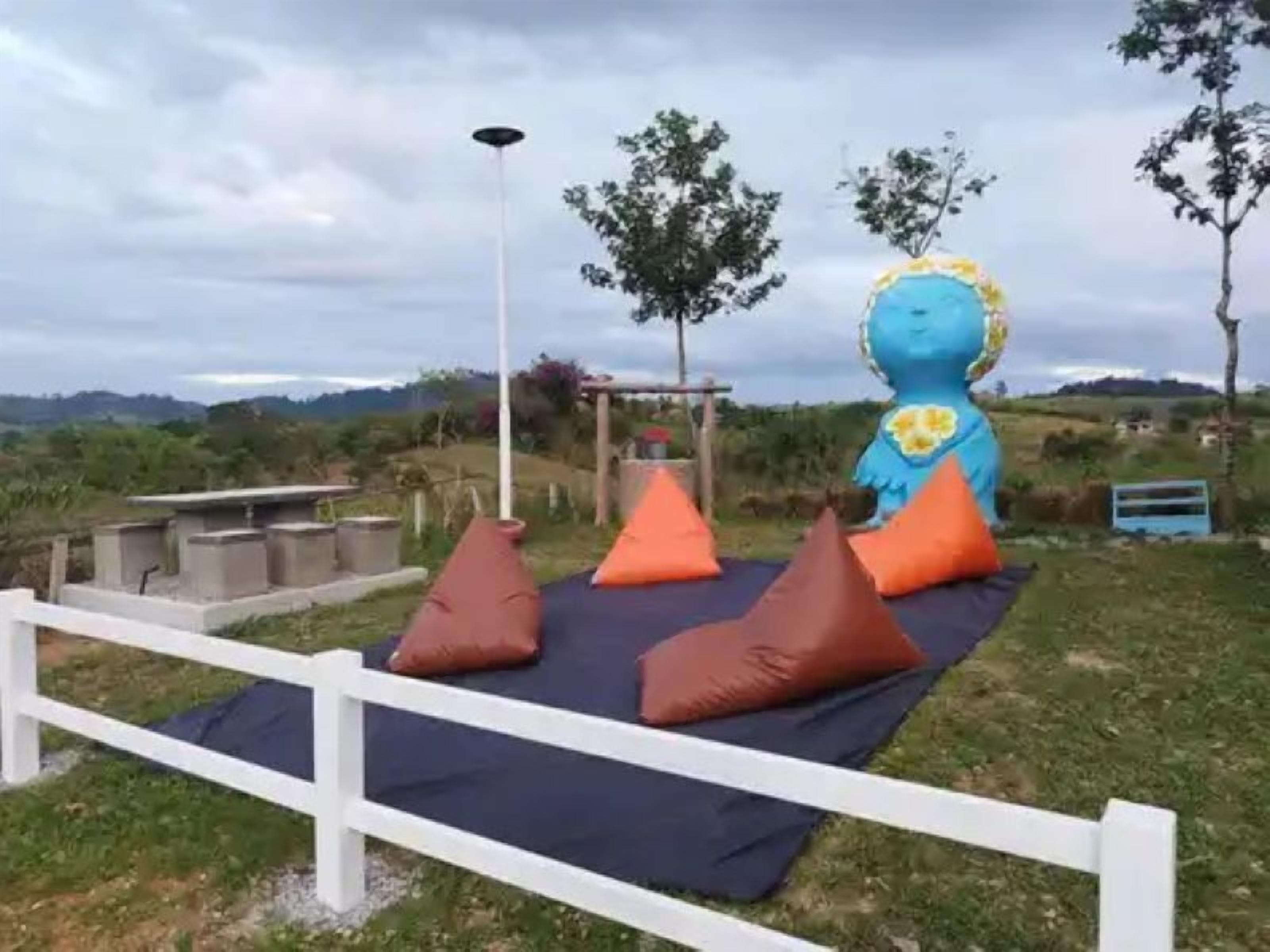 A blue inflatable sculpture with a sun on its head, surrounded by a white fence and orange cushions on a grassy area.
