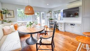 A modern kitchen with a dining area, featuring a round wooden table, black chairs, and a white sofa with orange pillows. - Hermes Haven Beach House - w/ Hot Tub, Blocks to beach (Encinitas)
