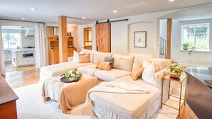 A spacious living area with a large L-shaped sofa, a coffee table, and a side table with a plant. A kitchen is visible in the background, and there is a sliding door leading to another room. - Hermes Haven Beach House - w/ Hot Tub, Blocks to beach (Encinitas)