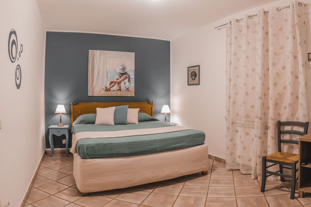 Ein Schlafzimmer mit einem Bett, zwei Nachttischen, einem Sessel, einem Bild an der Wand und einem Vorhang.