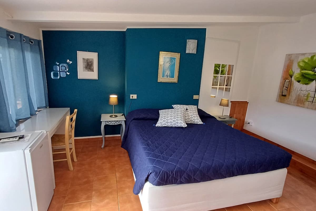 Ein kleines Zimmer mit Bett, Schreibtisch, Stuhl und einem kleinen Tisch mit Lampe.