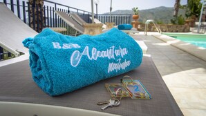 Toalla azul con el texto "B&B Alcantara Nativa" sobre una silla de descanso, llaves y un marco de fotos junto a una piscina con vistas a la montaña.