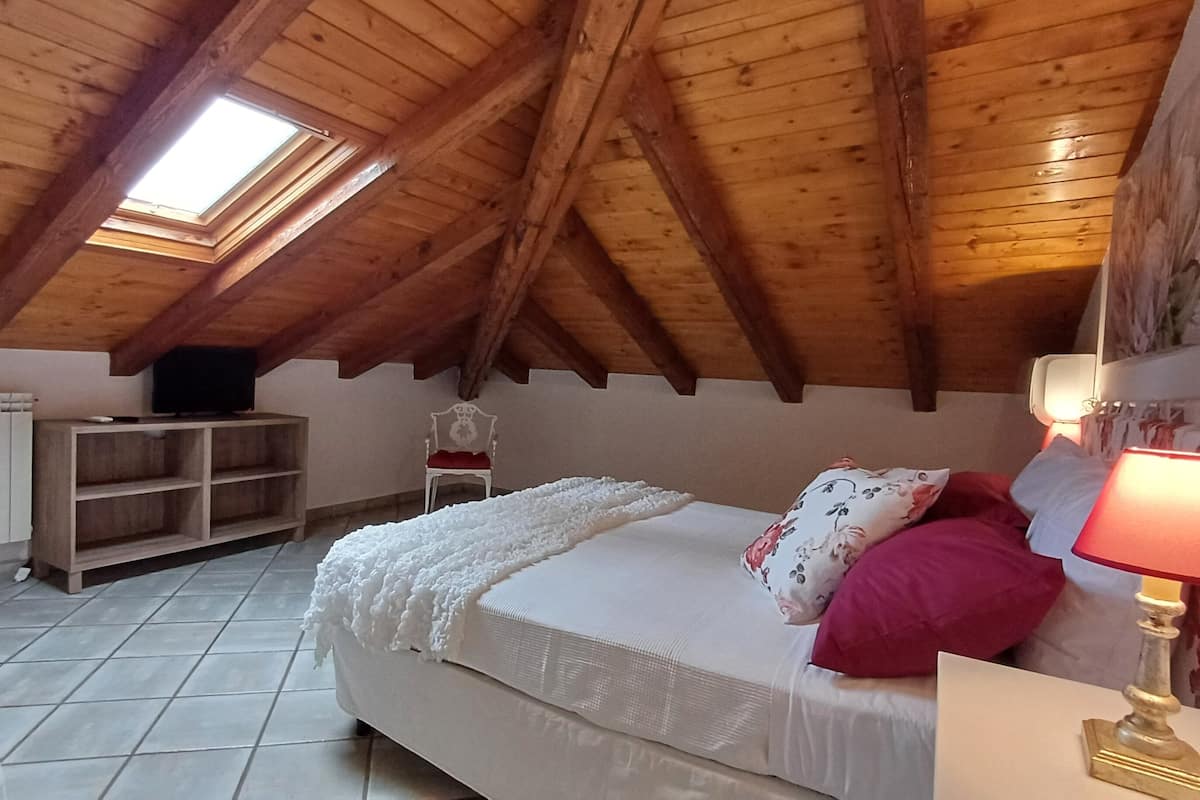 Ein Schlafzimmer mit schrägverlaufender Holzbalkendecke, einem Bett mit weißer Bettwäsche und rosa Kissen, einem Nachttisch mit Lampe, einem Heizkörper und einem Fernseher auf einem Regal.