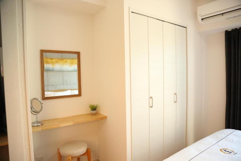 A bedroom with a bed, a wardrobe, a small desk with a mirror, and a shelf with a plant.