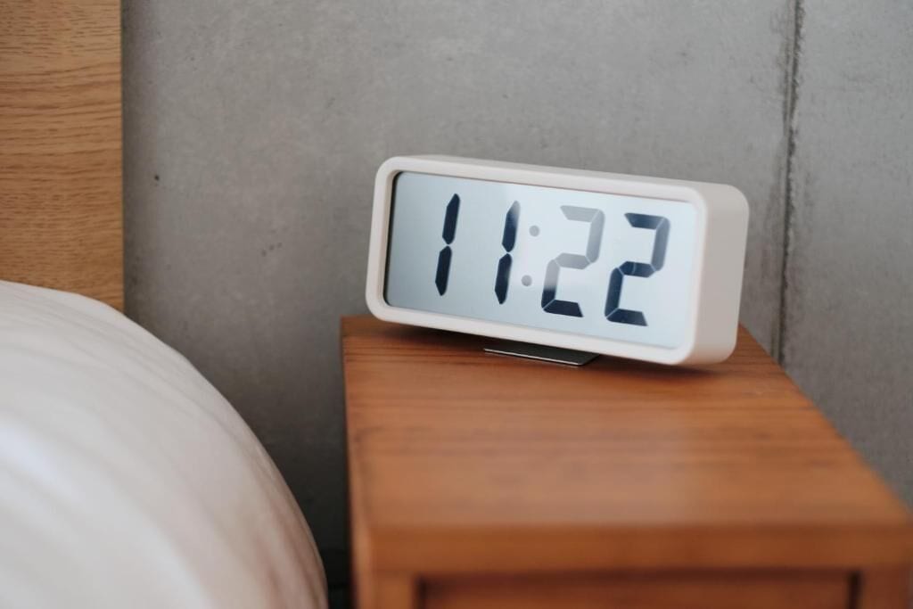 Un reloj digital muestra 11:22 sobre una mesita de noche de madera.