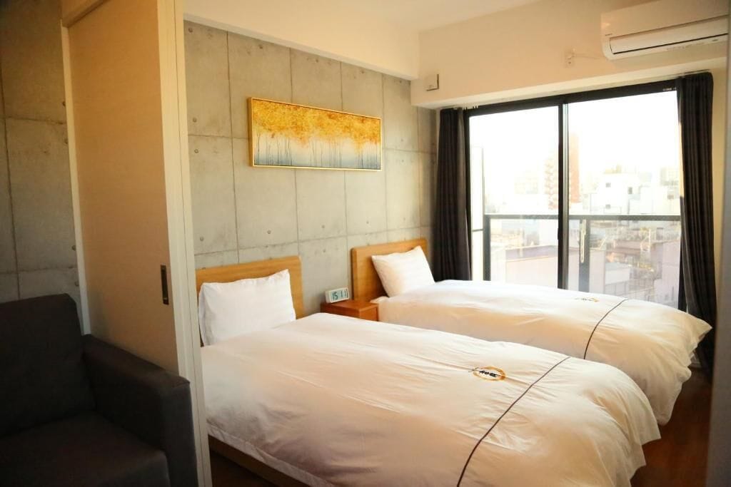 A hotel room with two beds, a large window with curtains, a painting on the wall, and a chair.