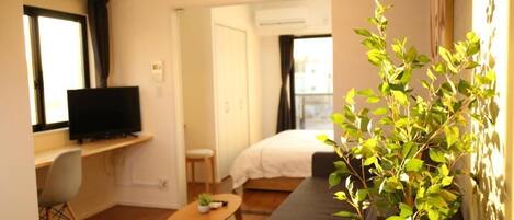 A compact hotel room with a bed, a TV on a desk, a small table, and a potted plant.