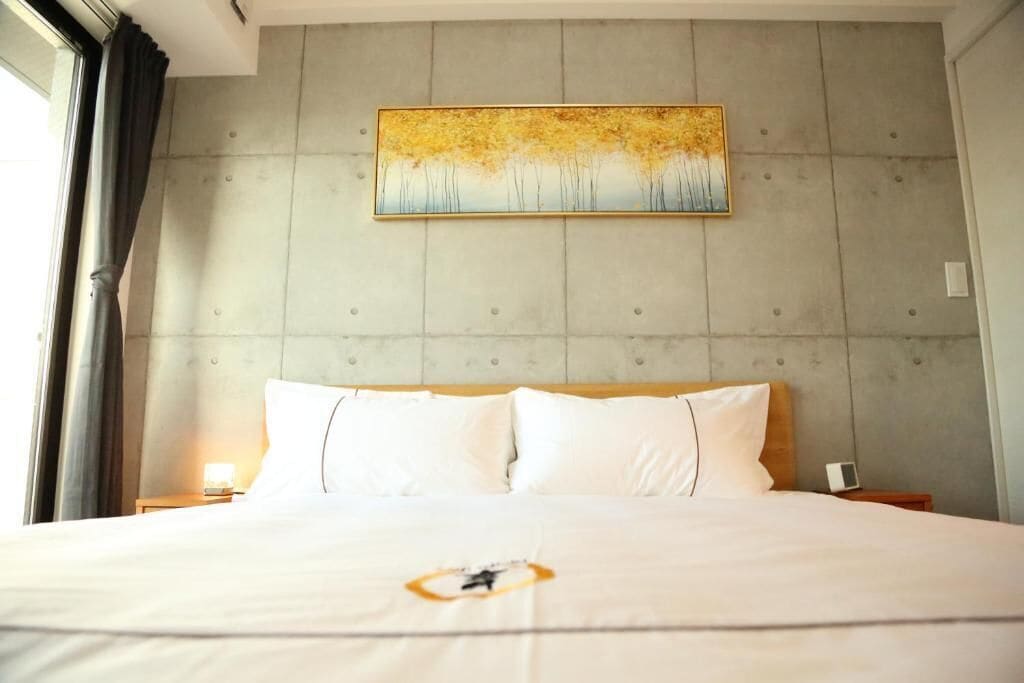 A hotel room with a large bed, a headboard with a textured wall, a painting above the bed, and a window with curtains.