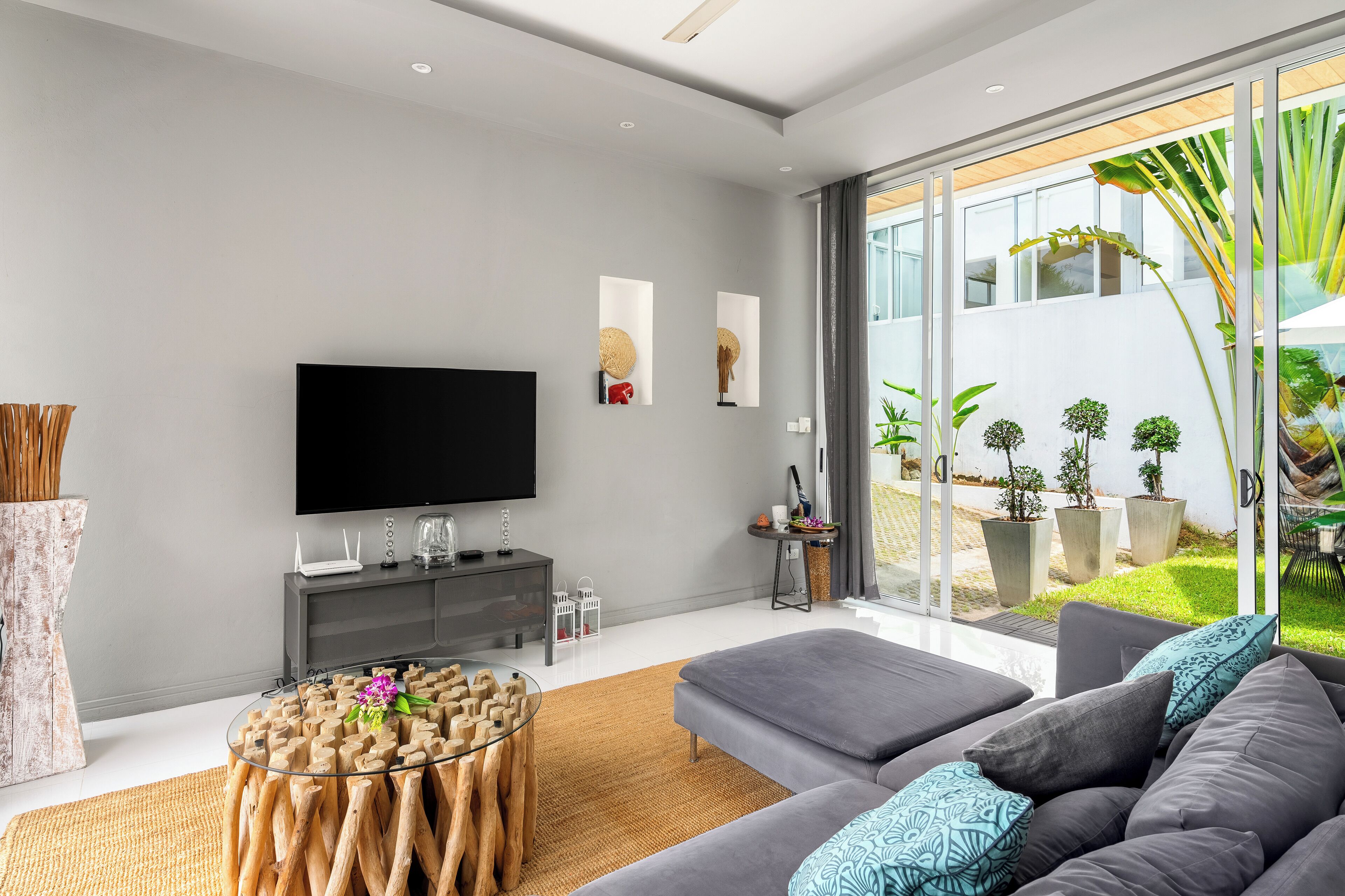 Un salon moderne doté d’un grand téléviseur à écran plat, de canapés gris, d’une table basse en verre et offrant une vue sur un jardin grâce à de grandes fenêtres.
