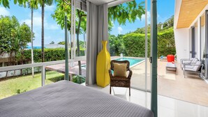 A modern bedroom with a large bed, a yellow vase, and a wicker chair. It overlooks a pool and lush greenery.