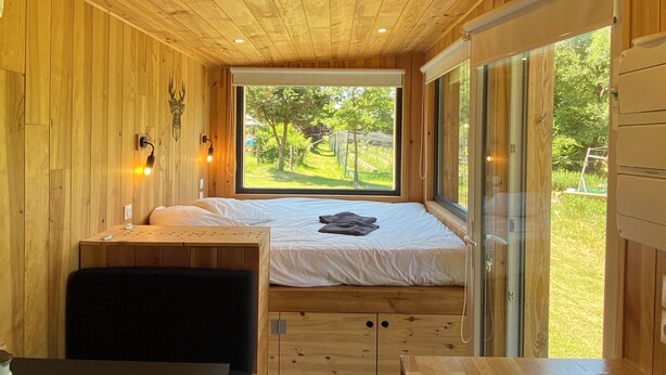 A wooden cabin room with a bed, a small table, and a bench. There is a window providing a view of the outdoors. - Lescargoterie de la Forge - Immersion Champêtre (Uzemain)