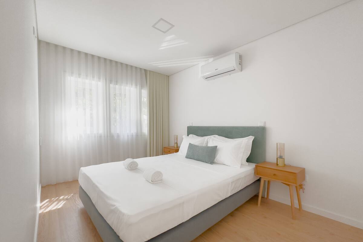 Une chambre à coucher moderne avec un grand lit, du linge de lit blanc, une tête de lit grise et un appareil de climatisation fixé au mur.
