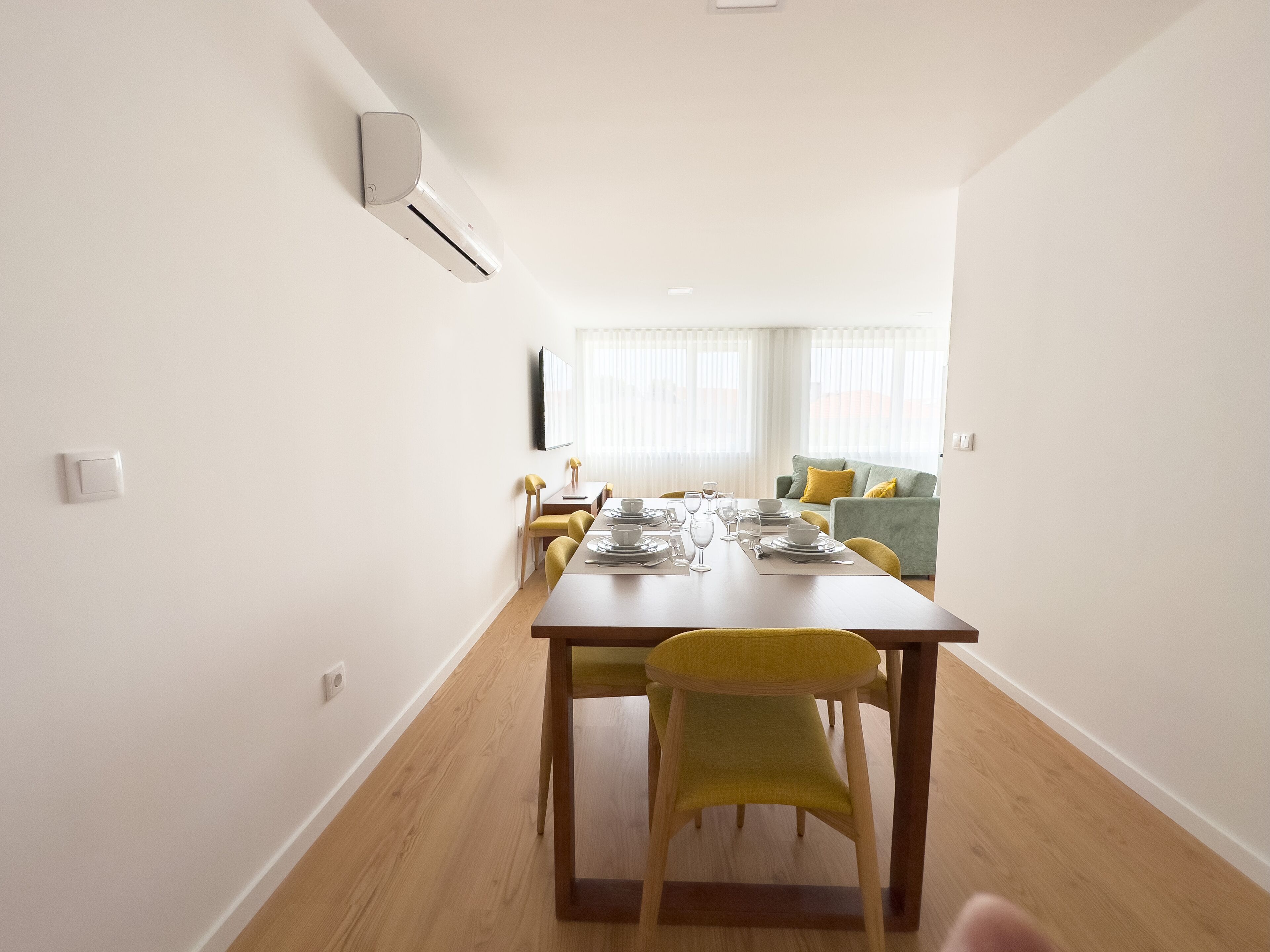 Un comedor moderno con una mesa de madera dispuesta para cuatro, sillas amarillas, un sofá verde y un equipo de aire acondicionado blanco en la pared.