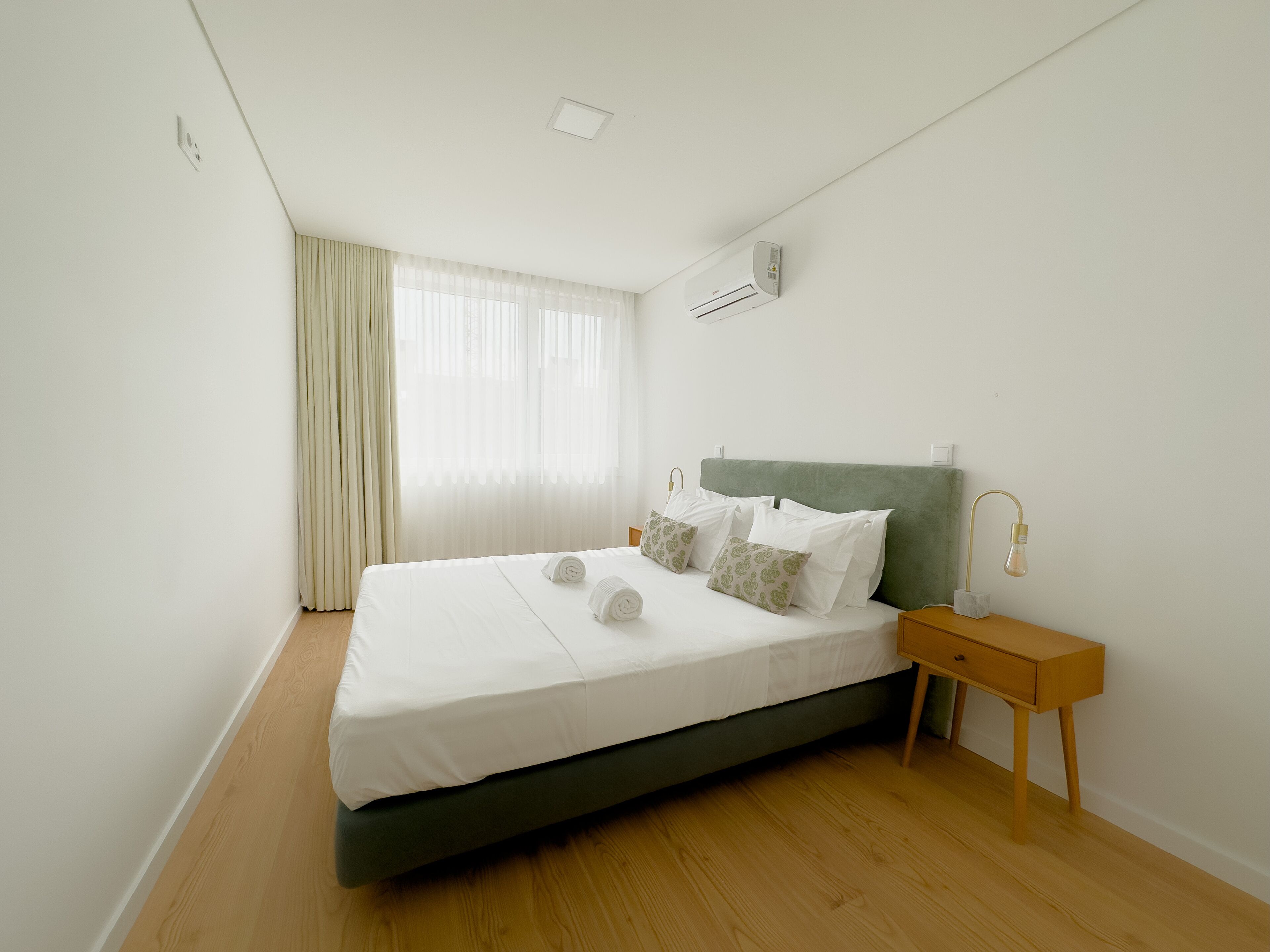 Una cama bien tendida con sábanas blancas y dos toallas enrolladas, una mesita de noche con una lámpara, y una ventana con cortinas traslúcidas.