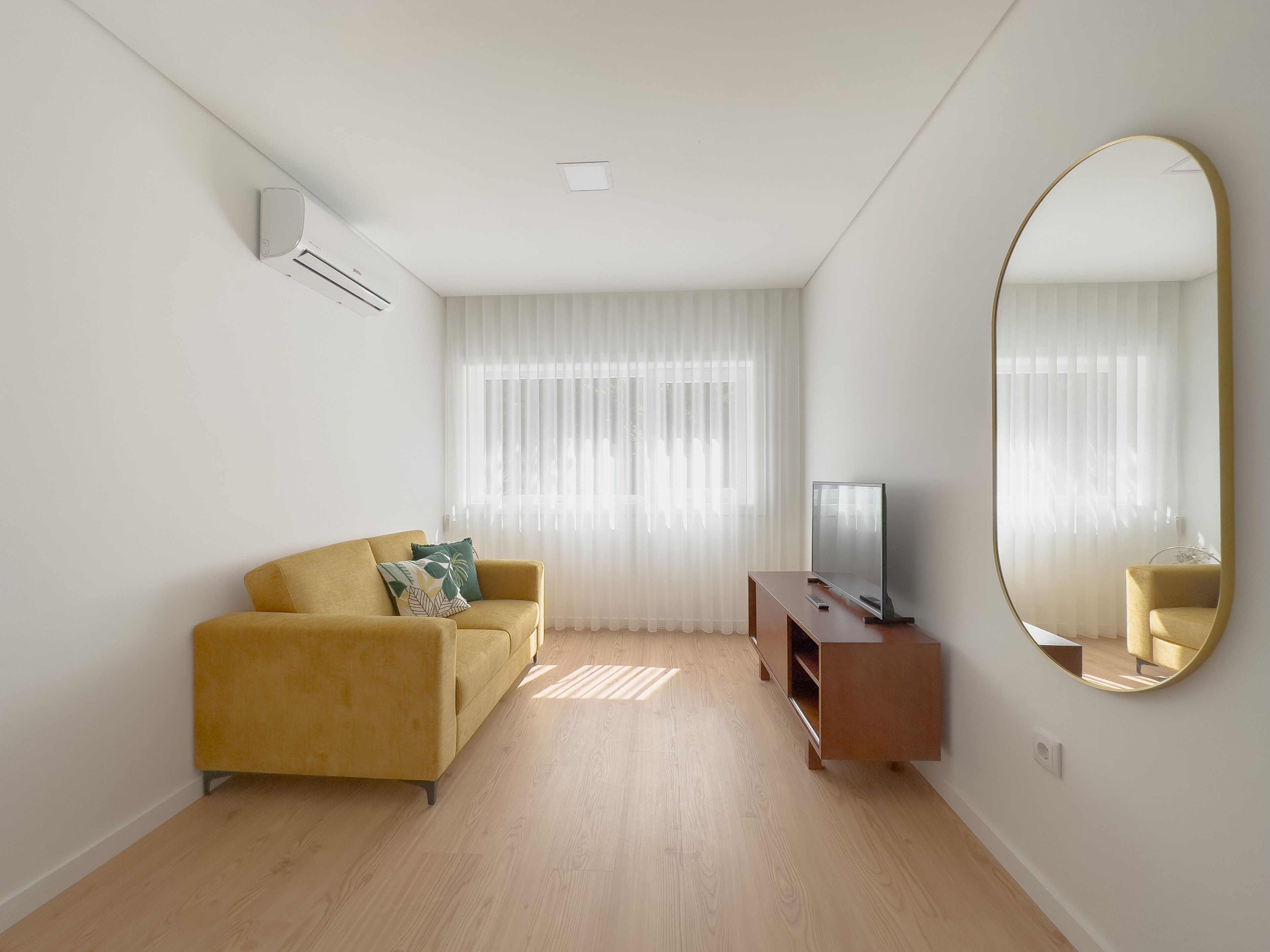 A modern living room with a yellow sofa, a flat-screen TV on a wooden stand, and a large mirror on the wall.