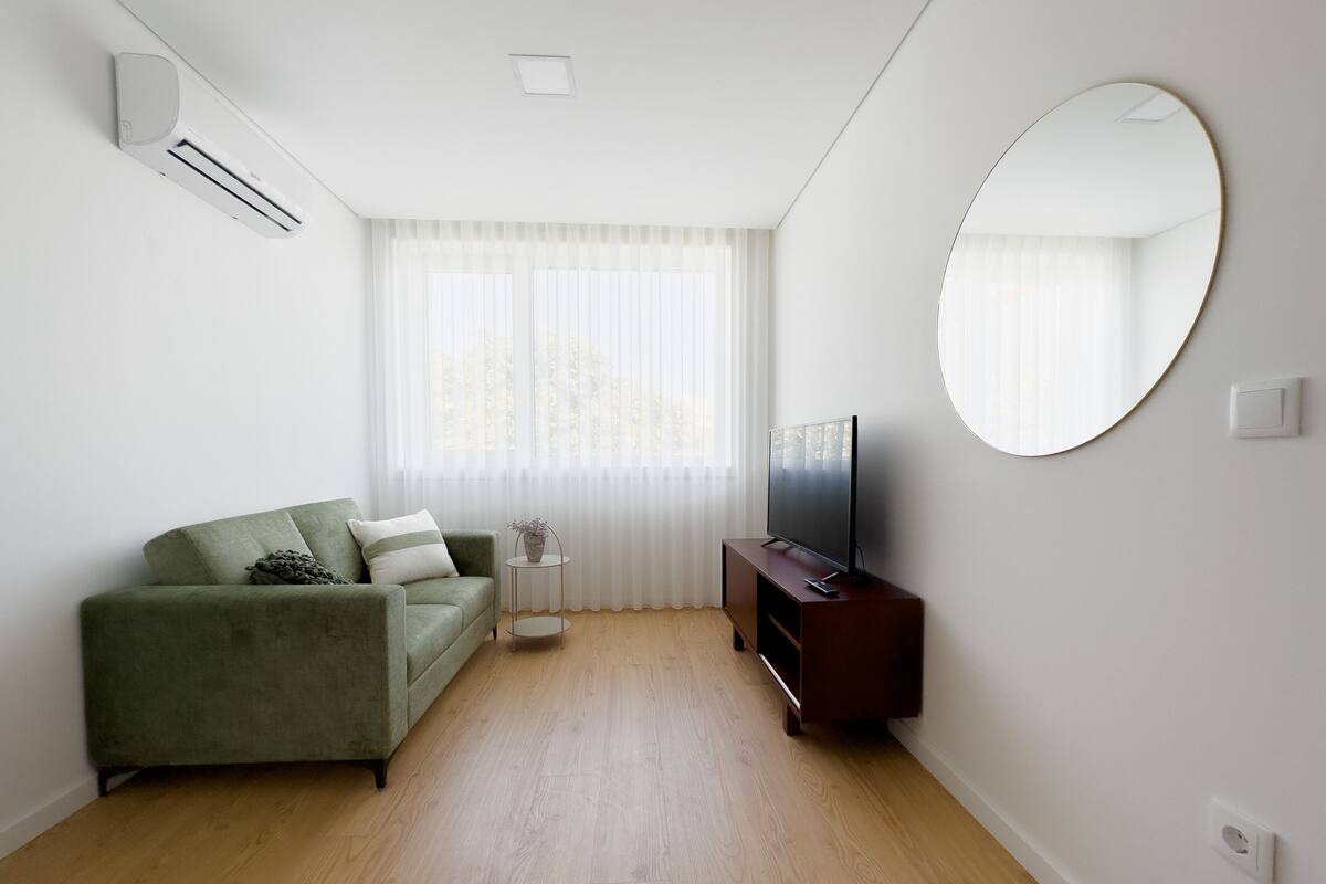 Un salon moderne avec un canapé vert, un téléviseur à écran plat posé sur un meuble en bois et un grand miroir rond accroché au mur.