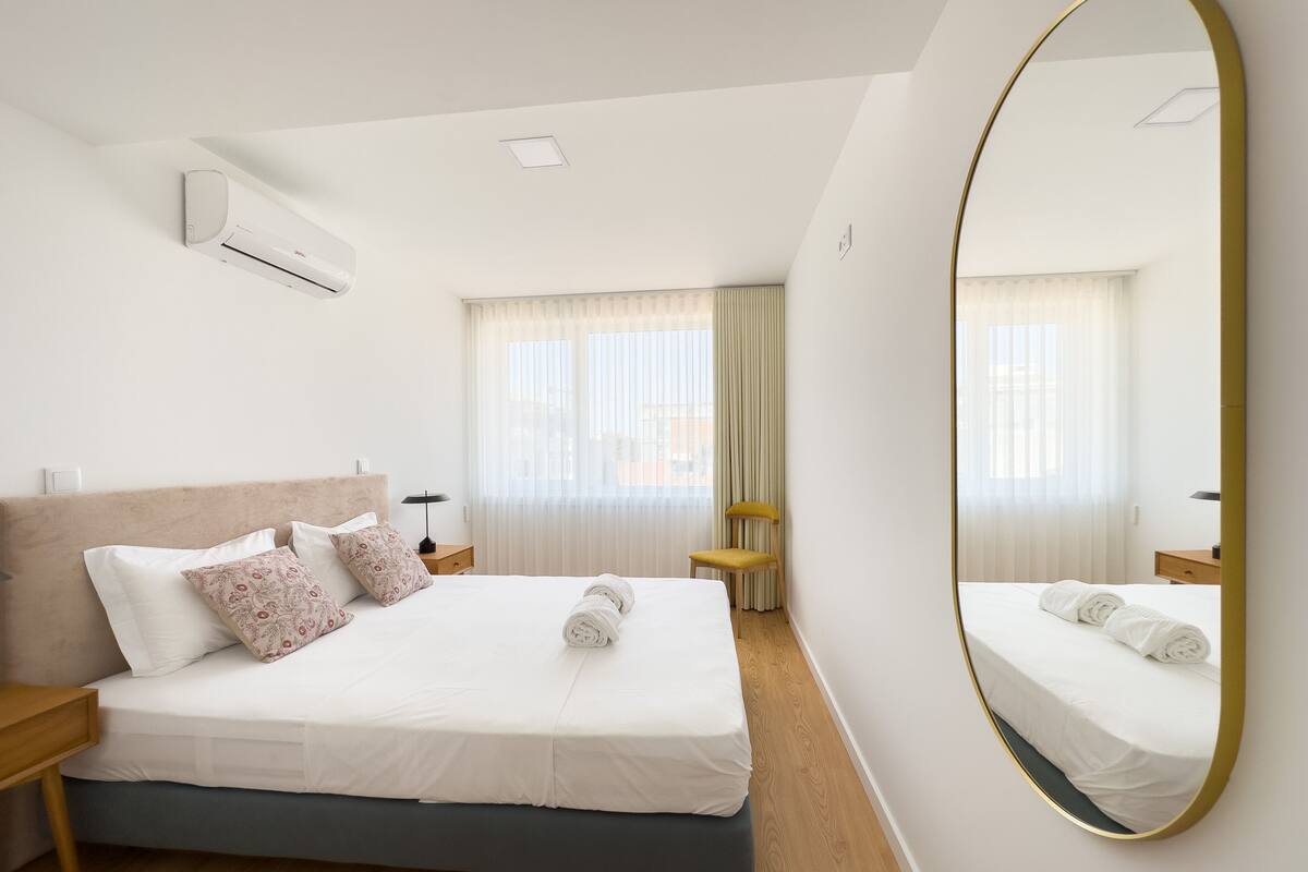 Une chambre d’hôtel moderne avec un grand lit, un miroir rond et une fenêtre avec des rideaux légers.