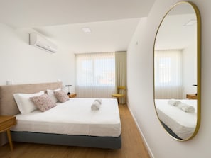 Une chambre d’hôtel moderne avec un grand lit, un miroir rond et une fenêtre avec des rideaux légers.