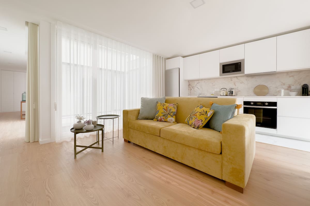 Una sala de estar moderna con un sofá amarillo, una mesita auxiliar y una cocina con armarios blancos y encimera de mármol.